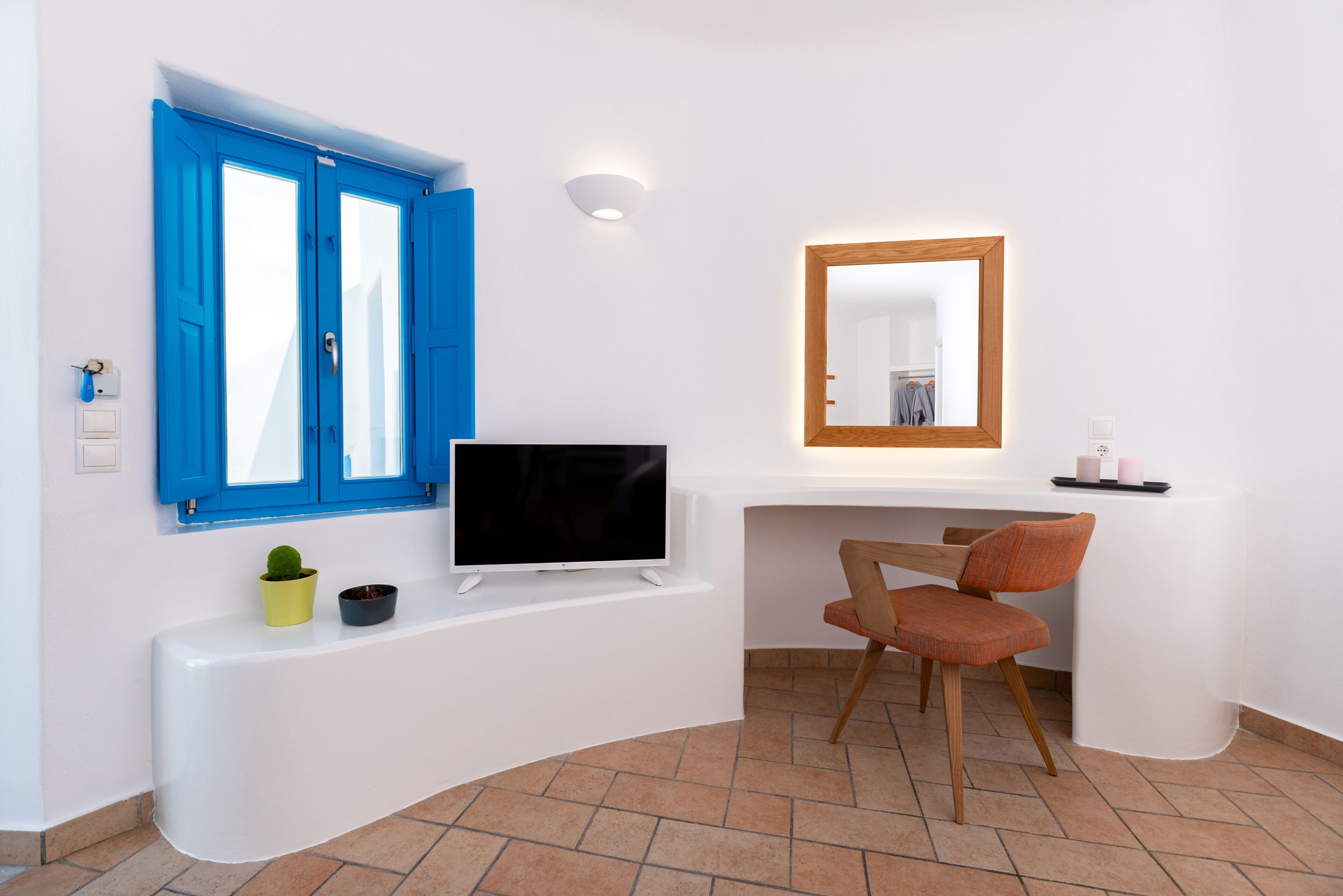 A modern room with a white desk, a flat-screen TV, a chair, a small potted plant, and a blue-framed window.