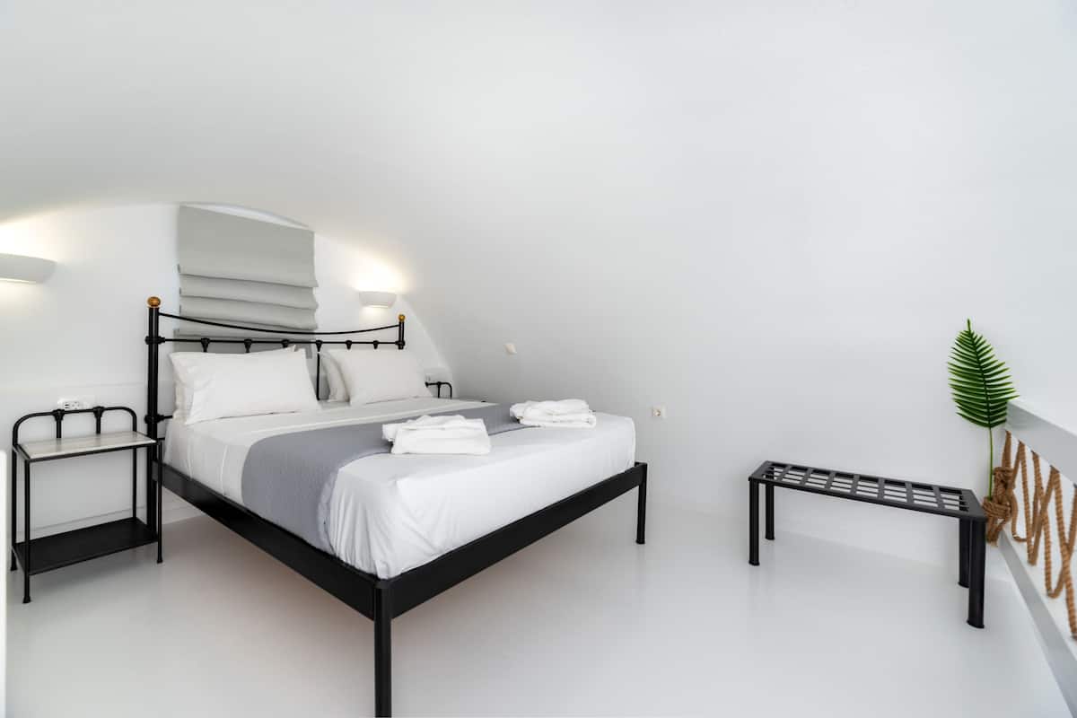A modern bedroom with a black metal bed frame, white bedding, a bedside table with a lamp, and a small bench.