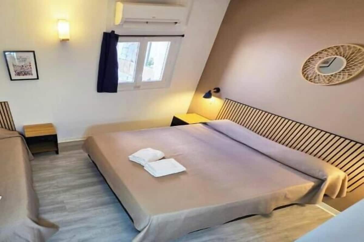 A small, compact room with two single beds, a bedside table, a wall-mounted air conditioner, a window with curtains, and a framed picture on the wall.