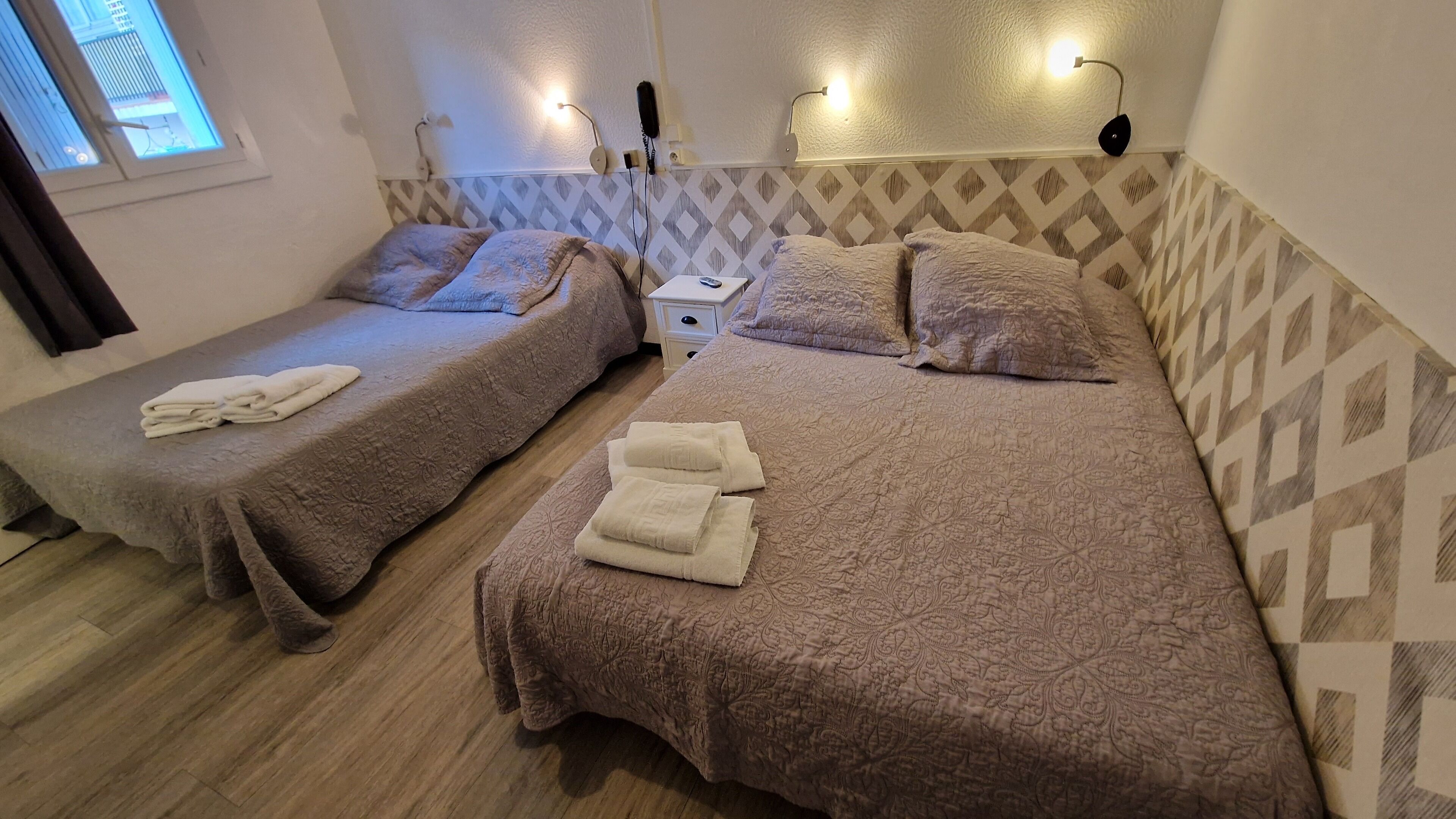 Una habitación de hotel con dos camas, cada una con una colcha y almohadas estampadas, una mesita de noche con lámpara y una ventana con persianas.