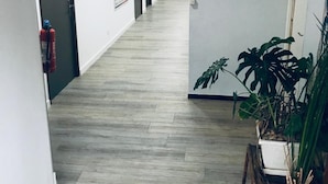 Un pasillo con piso de madera clara, paredes blancas y una puerta a la izquierda. Hay plantas en macetas a la derecha.