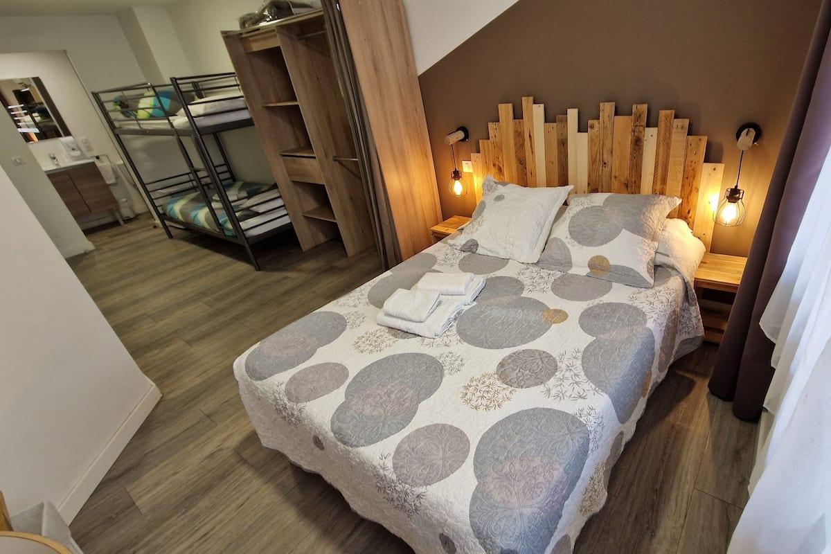 A bedroom with a bed, wooden headboard, bedside lamps, and a bunk bed in the background.