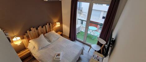 A bedroom with a bed, bedside lamps, a window with curtains, a small table with a plant, and a view of a balcony with outdoor furniture.