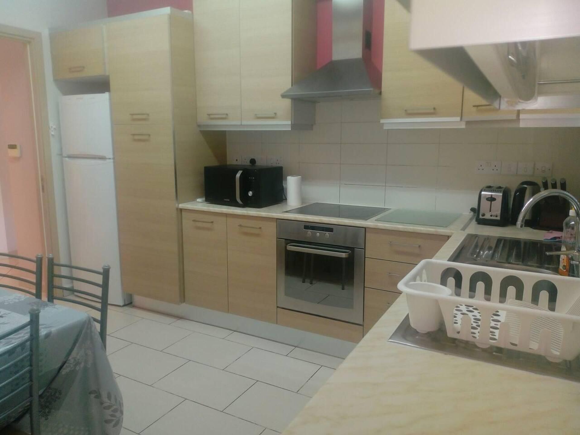 Cucina compatta con mobili chiari in legno, forno in acciaio inossidabile, microonde e tostapane. Si nota un lavandino piccolo con scolapiatti e un tavolo con sedie in primo piano.