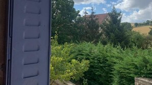 Una vista attraverso una finestra con inferriata, su un paesaggio con alberi e un edificio all'orizzonte, sotto un cielo parzialmente nuvoloso.