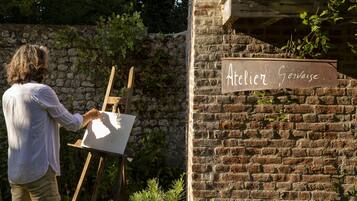 Une personne est en train de peindre sur un chevalet devant un mur de briques sur lequel est apposée une enseigne indiquant « Atelier Gevaise ».