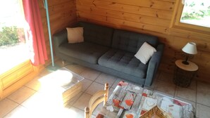 Ein gemütliches Zimmer mit grauem Sofa, zwei Kissen, einem kleinen Tisch mit Lampe und einem Fenster mit Vorhängen.