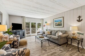 A living room with a sofa, a coffee table, and a television. There is a balcony with a view of the outdoors.