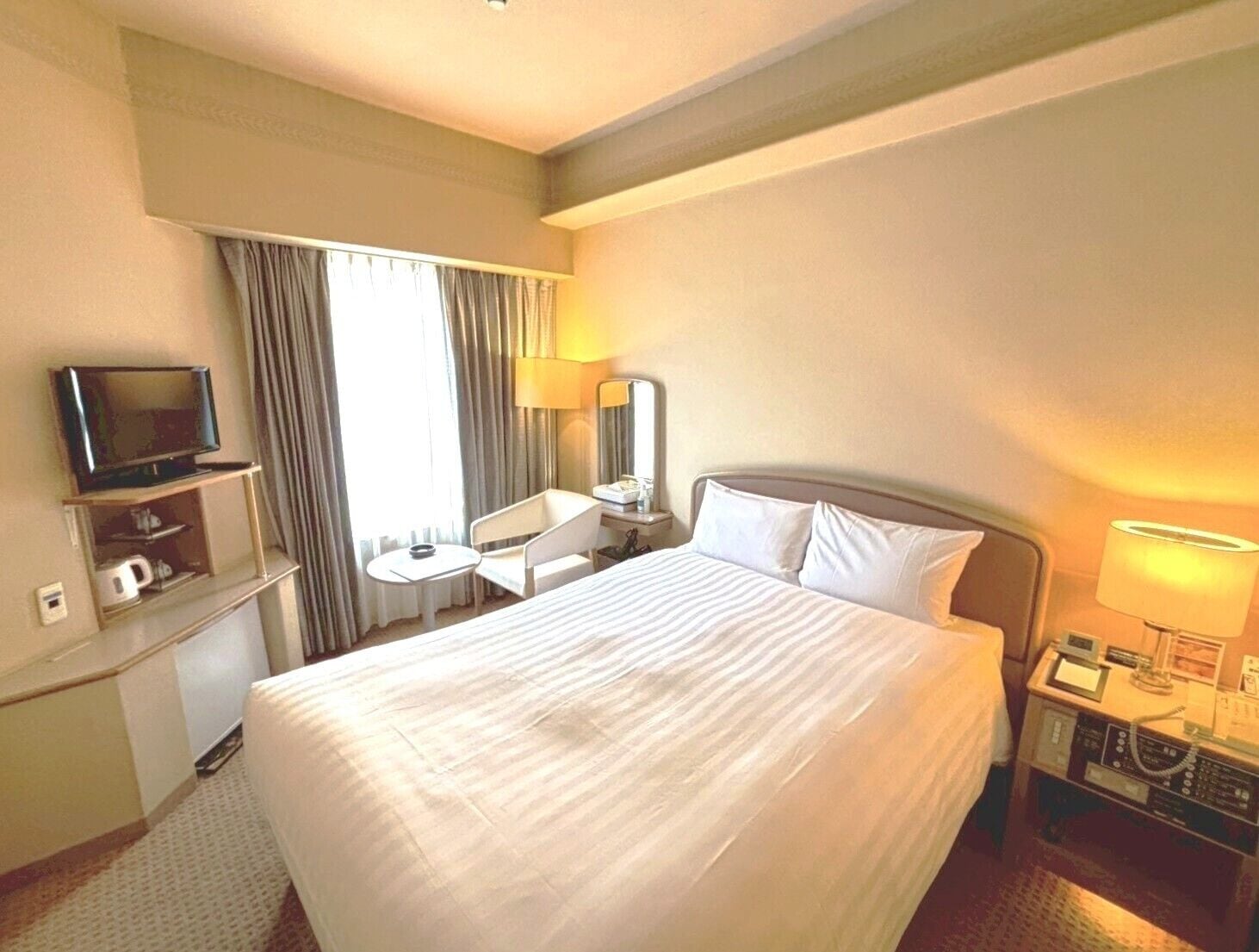 A hotel room with a bed, a TV on a stand, a desk with a chair, a lamp, and a telephone.