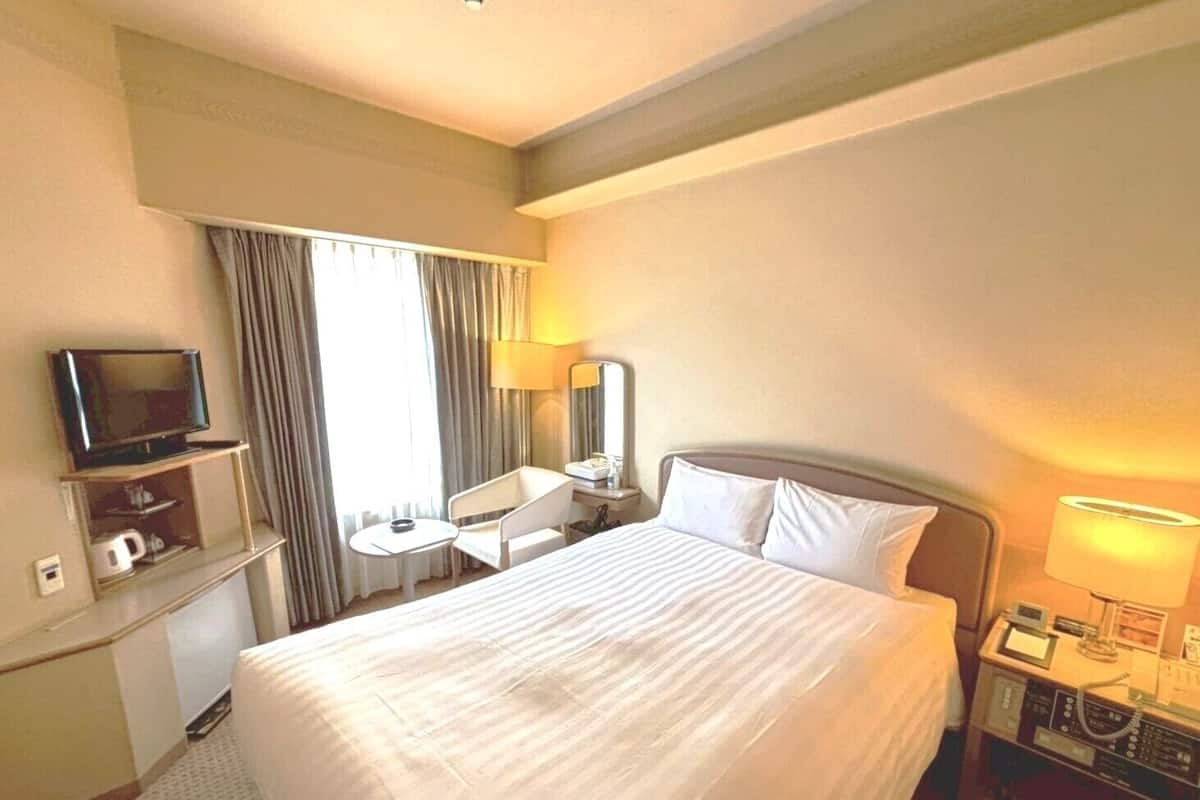 Une chambre d’hôtel comprenant un lit, un téléviseur posé sur un meuble, un bureau avec une chaise, une lampe et un téléphone.