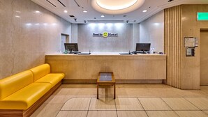 Un área de recepción con mostrador, un sofá amarillo, una mesita y un cartel en la pared que dice 'Smile Hotel'.