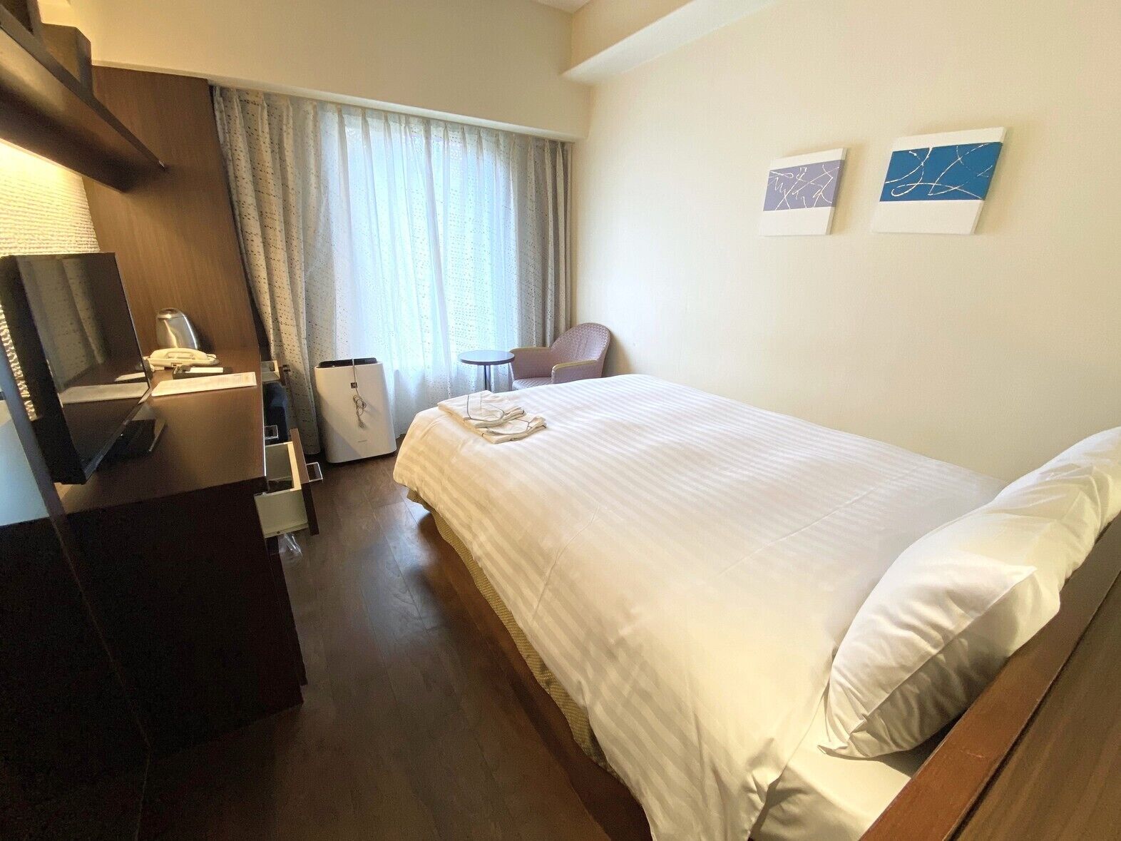 A hotel room with a bed, a desk with a telephone, a chair, a small table, and a wall-mounted TV.