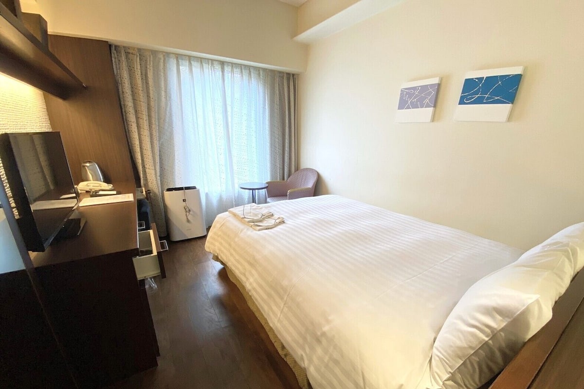 Habitación de hotel con cama, escritorio con teléfono, silla, mesita pequeña y televisor montado en la pared.