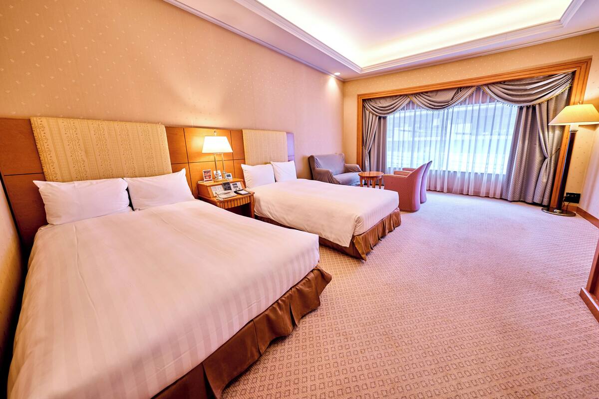 A hotel room with two beds, a desk, a chair, and a large window with curtains.