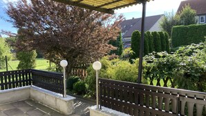 Een overdekt terras met stenen vloer, metalen hekwerk en uitzicht op een tuin met verschillende planten en een boom.