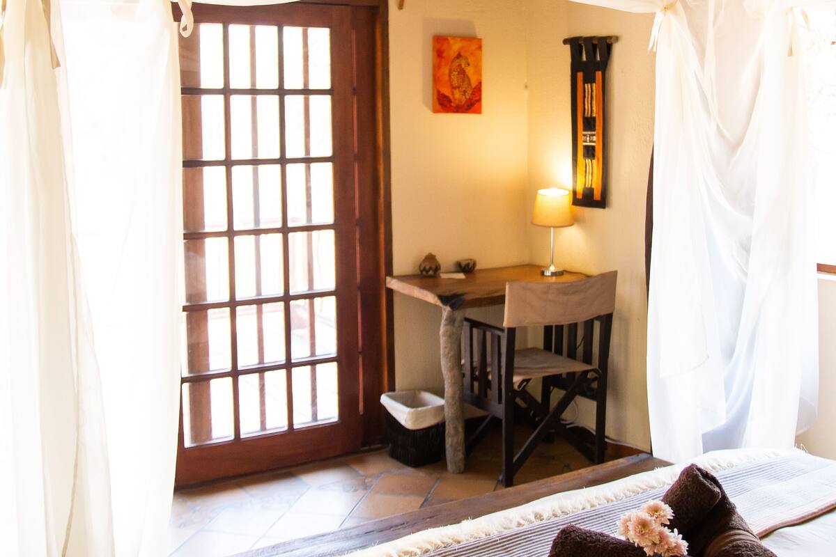 A room with a bed, a desk, and a chair. There is a window with bars and a door.