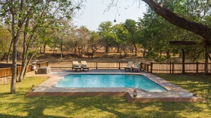 Une piscine rectangulaire avec une terrasse carrelée, entourée d’une clôture en bois et d’arbres.