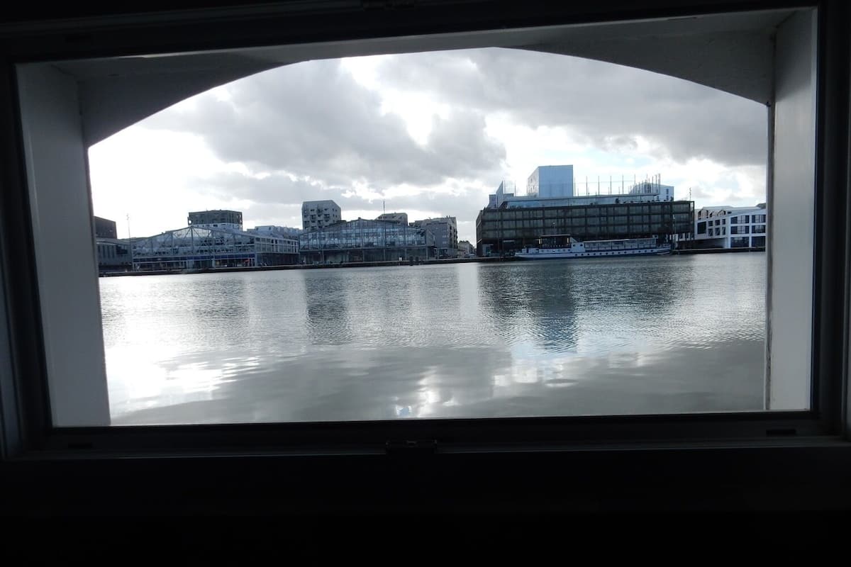 Une vue sur un paysage urbain à travers une fenêtre, avec un grand bâtiment à la façade vitrée et une étendue d'eau au premier plan.