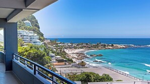 A coastal view from a balcony overlooking a beach with clear blue waters and rocky shoreline. - Clifton YOLO Spaces – Clifton Beachfront Executive  (Cape Town)