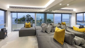 A modern living room with a grey sectional sofa, yellow cushions, and patterned pillows. It features large floor-to-ceiling windows offering a view of the ocean and a balcony. - Clifton YOLO Spaces – Clifton Beachfront Executive  (Cape Town)