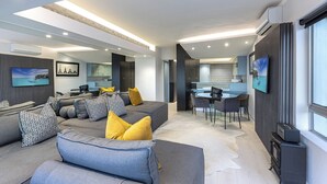 A modern living room with a grey sofa, yellow cushions, a dining area with a glass table, and a kitchen with dark cabinetry. - Clifton YOLO Spaces – Clifton Beachfront Executive  (Cape Town)