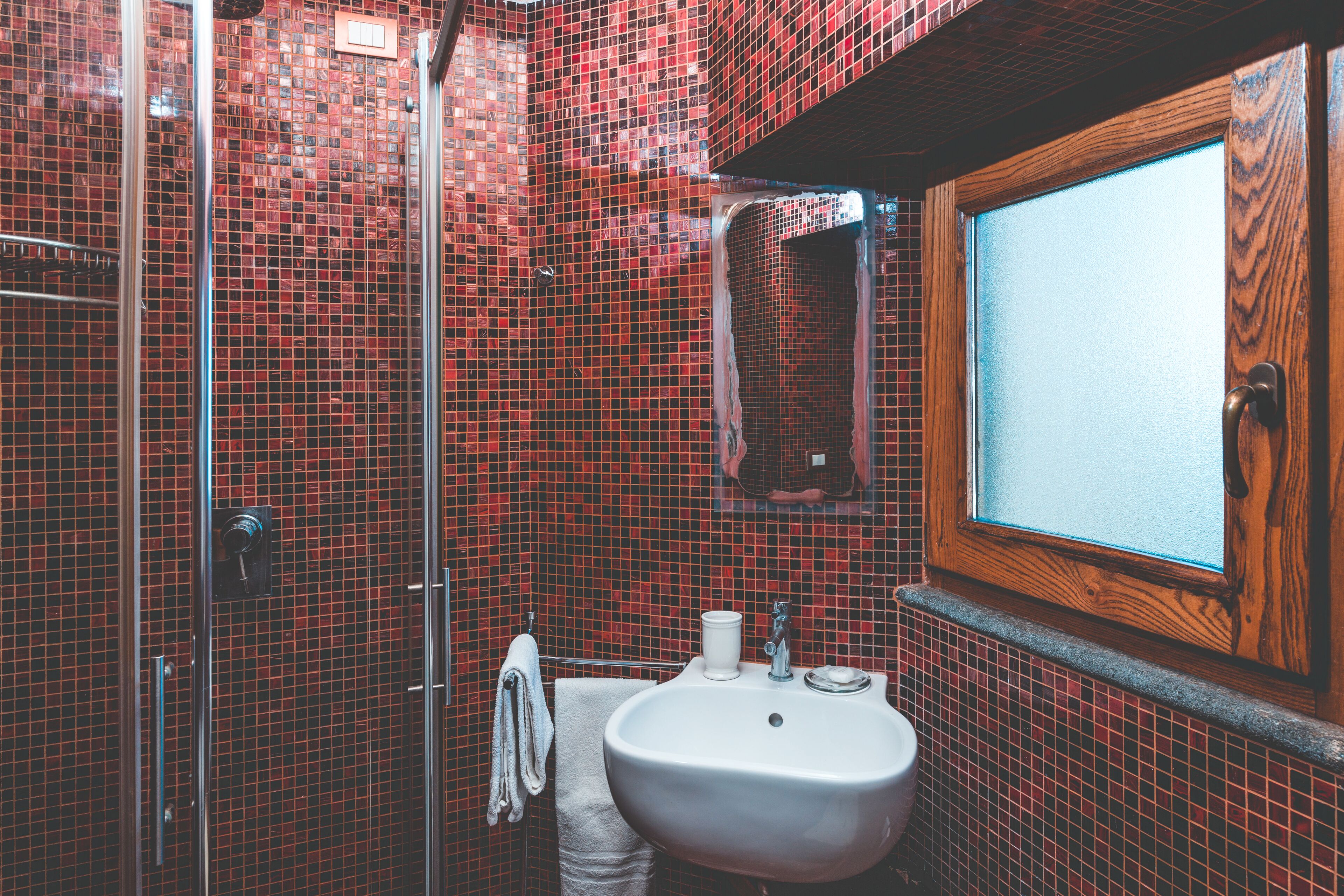 Ein Badezimmer mit roten Mosaikfliesen, einem Holzrahmenfenster, einem weißen Waschbecken und einer Duschkabine aus Glas.