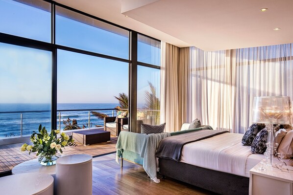 Ein modernes Schlafzimmer mit großem Fenster mit Meerblick, einem Bett mit vielen Kissen, einem Nachttisch und einer kleinen Sitzecke mit Pflanze.