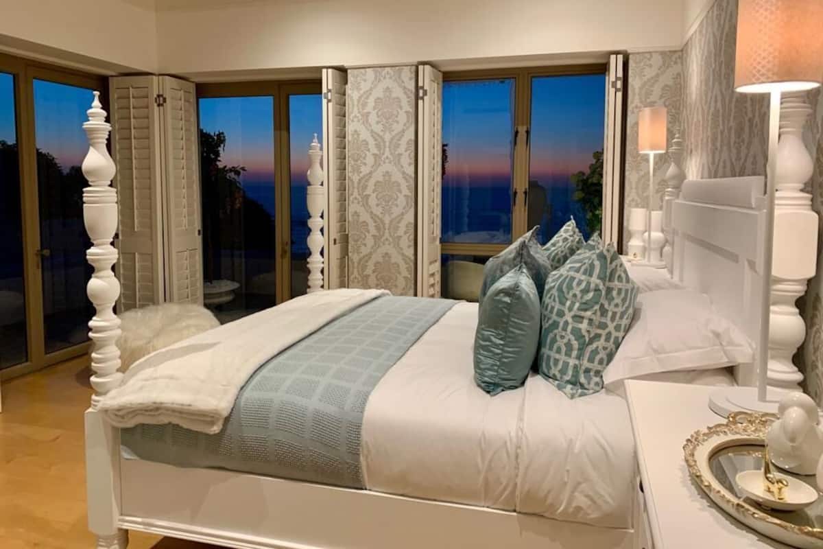 A bedroom with a bed, a nightstand, a lamp, and a view of the sunset through large windows.