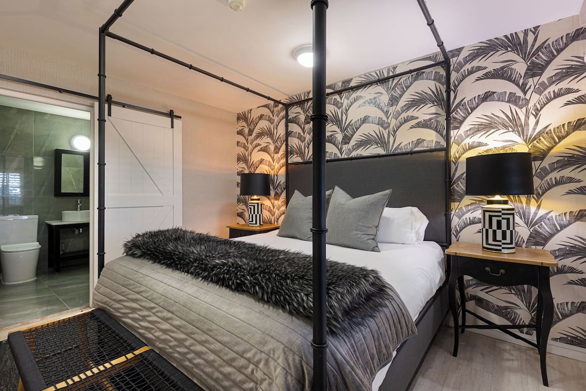 A modern bedroom with a four-poster bed, a large mirror, and a tropical wallpaper design.