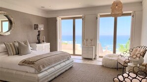 A bedroom with a large bed, a nightstand, a dresser, a chair, and a view of the ocean. - Clifton YOLO Spaces – Clifton Mansion Villa (Cape Town)
