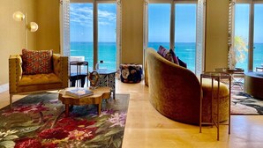 A spacious living room with a large window offering a view of the ocean, a plush armchair, a coffee table, and a small side table. - Clifton YOLO Spaces – Clifton Mansion Villa (Cape Town)