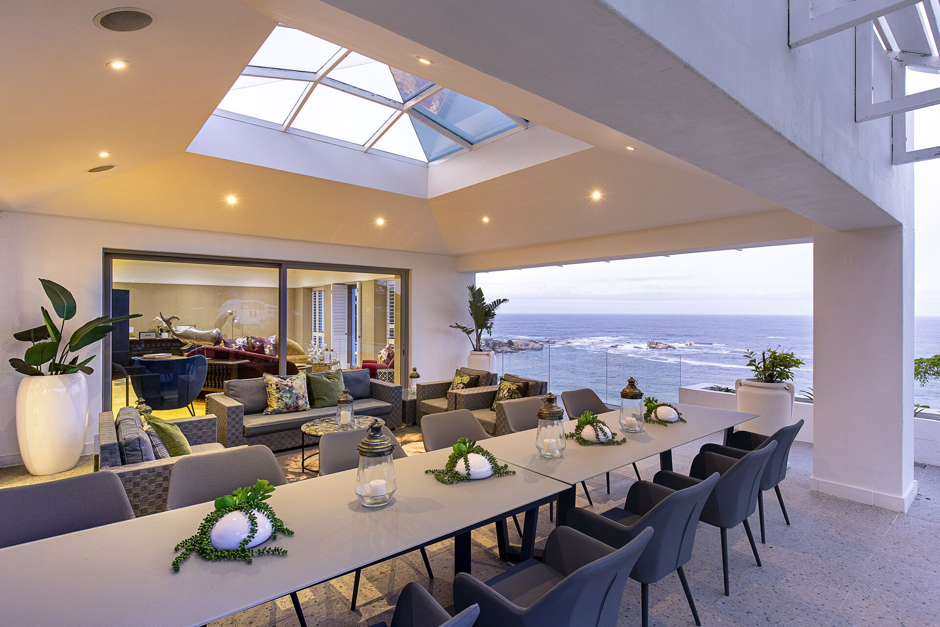 A modern dining area with a view of the ocean, featuring a long table with chairs, potted plants, and decorative items.