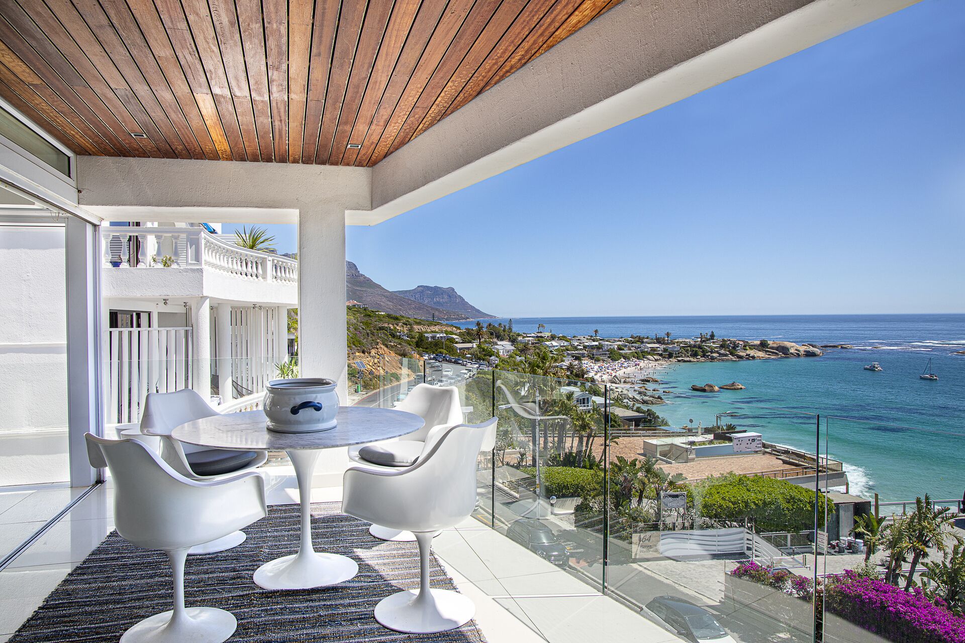 A balcony with a round table and four white chairs, overlooking a coastal town and the sea.