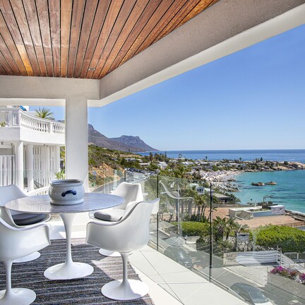 A balcony with a round table and four white chairs, overlooking a coastal town and the sea.