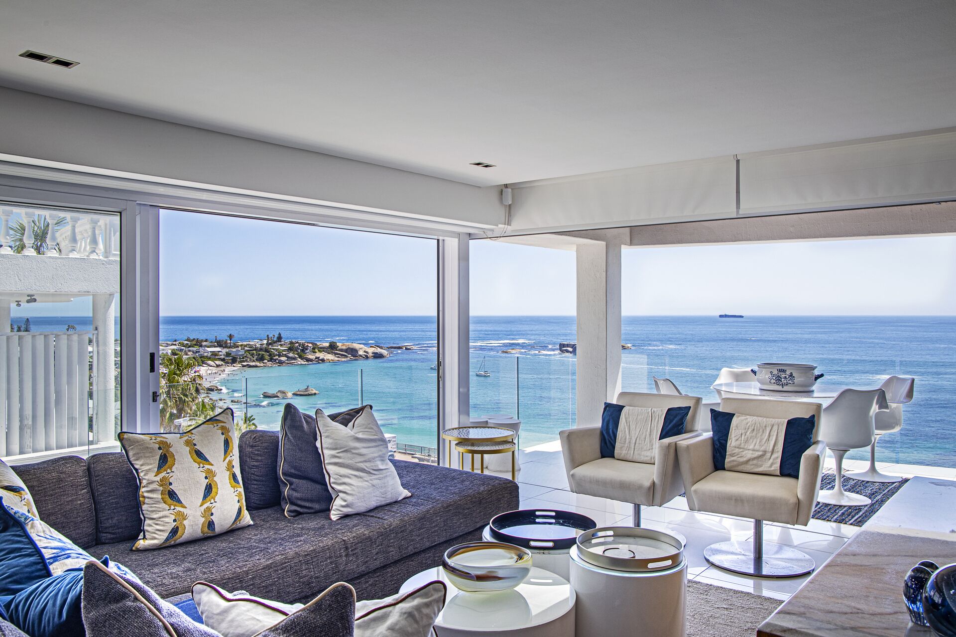 A modern living room with a large balcony overlooking the sea, featuring a sofa, chairs, and a dining table.
