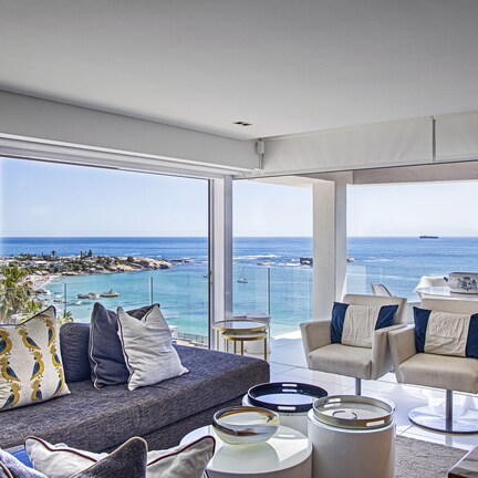 A modern living room with a large balcony overlooking the sea, featuring a sofa, chairs, and a dining table.