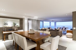 A modern dining area with a wooden table and white chairs, a sofa with cushions, and a view of the ocean through large windows.
