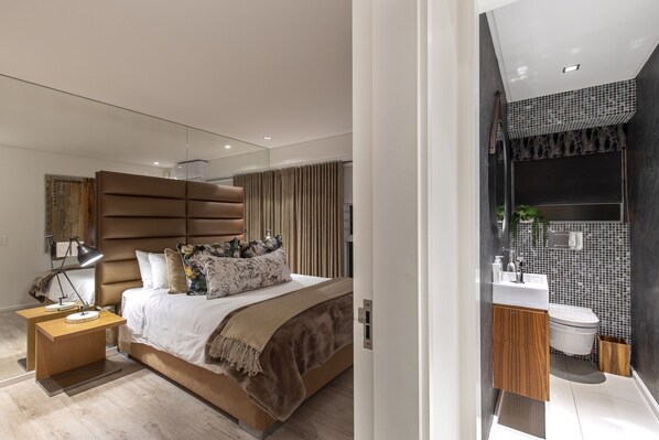 Ein modernes Schlafzimmer mit einem großen Bett, einem Nachttisch und einem Beistelltisch. Das Badezimmer verfügt über ein modernes Waschbecken, einen Spiegel und geflieste Wände.