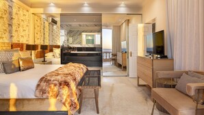 Ein geräumiges Hotelzimmer mit einem großen Bett, einer Sitzecke mit Sofa und Sessel, einem Fernseher und Blick auf das Meer durch eine geöffnete Tür.