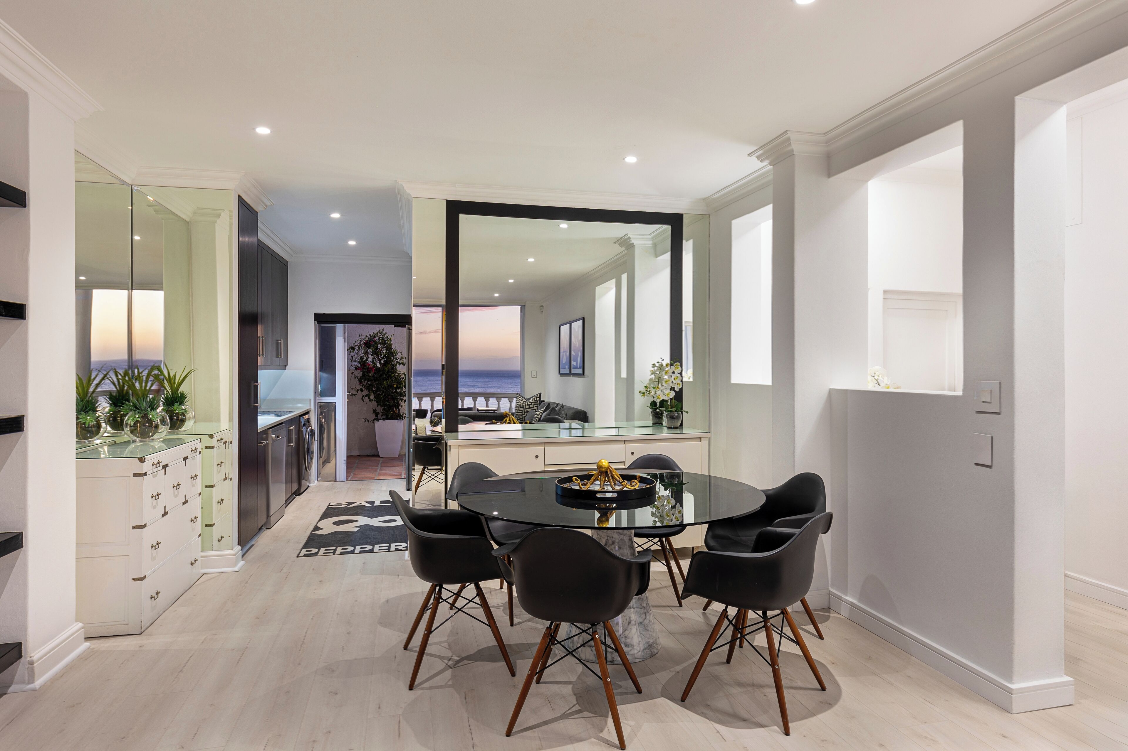 A modern dining area with a glass table, black chairs, and a view of the ocean through a large window.