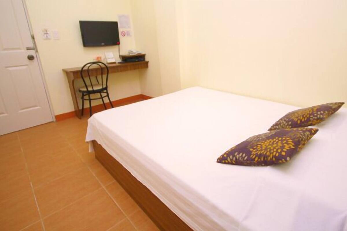 Un dormitorio con una cama, dos almohadas, un escritorio con una silla, un televisor en la pared y una puerta.
