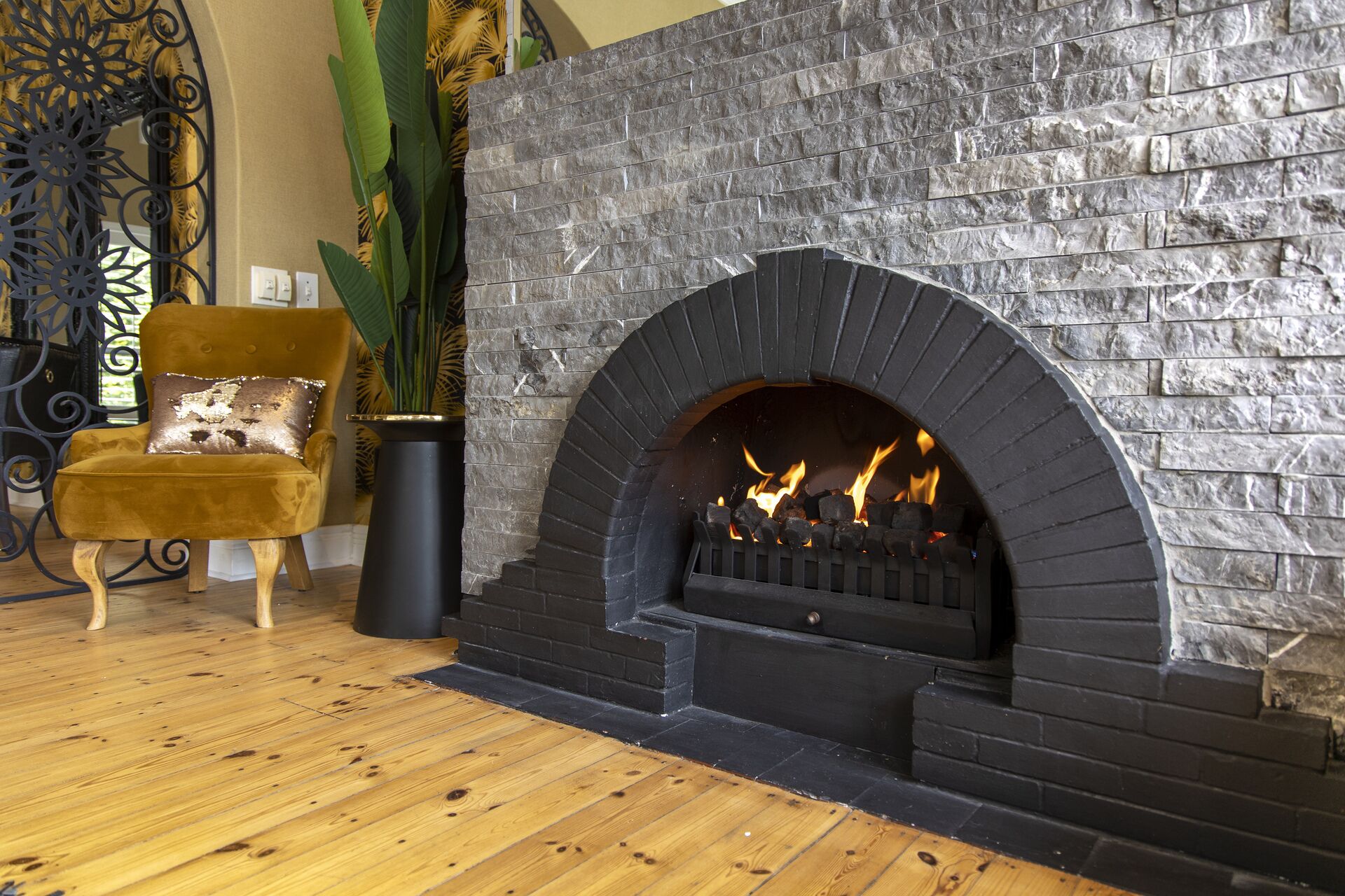 Una chimenea de piedra con fuego, un sillón cómodo y una planta en maceta en una habitación con suelo de madera.