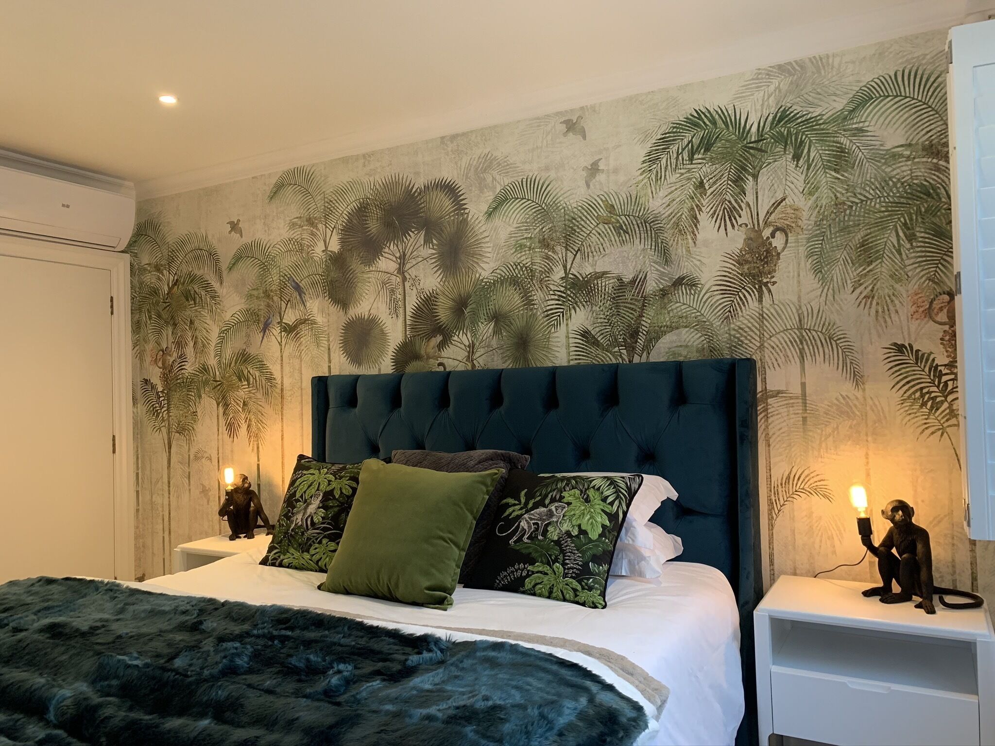 Een slaapkamer met een gestoffeerd hoofdeinde, een bed met een donkergroene deken, en een muurschildering van tropisch groen. Er staan twee nachtkastjes met lampen en een klein beeldje op een ervan.