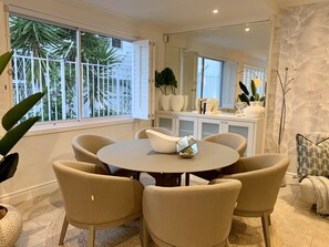 A dining area with a round table and six chairs, a large window with a view of palm trees, and a mirrored wall reflecting the room. - Clifton YOLO Spaces – Clifton Boutique Apartments  (Cape Town)