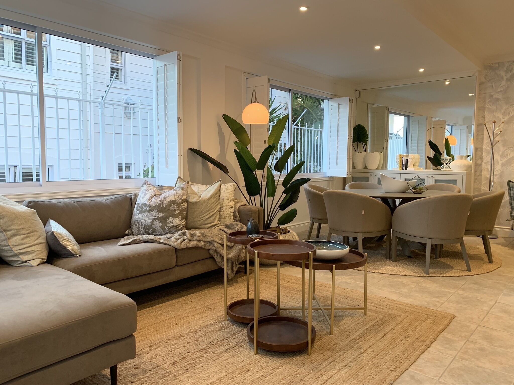 A modern living room with a sofa, coffee table, and chairs. There is a dining area with a table and chairs. Large windows provide natural light and views of the outside.