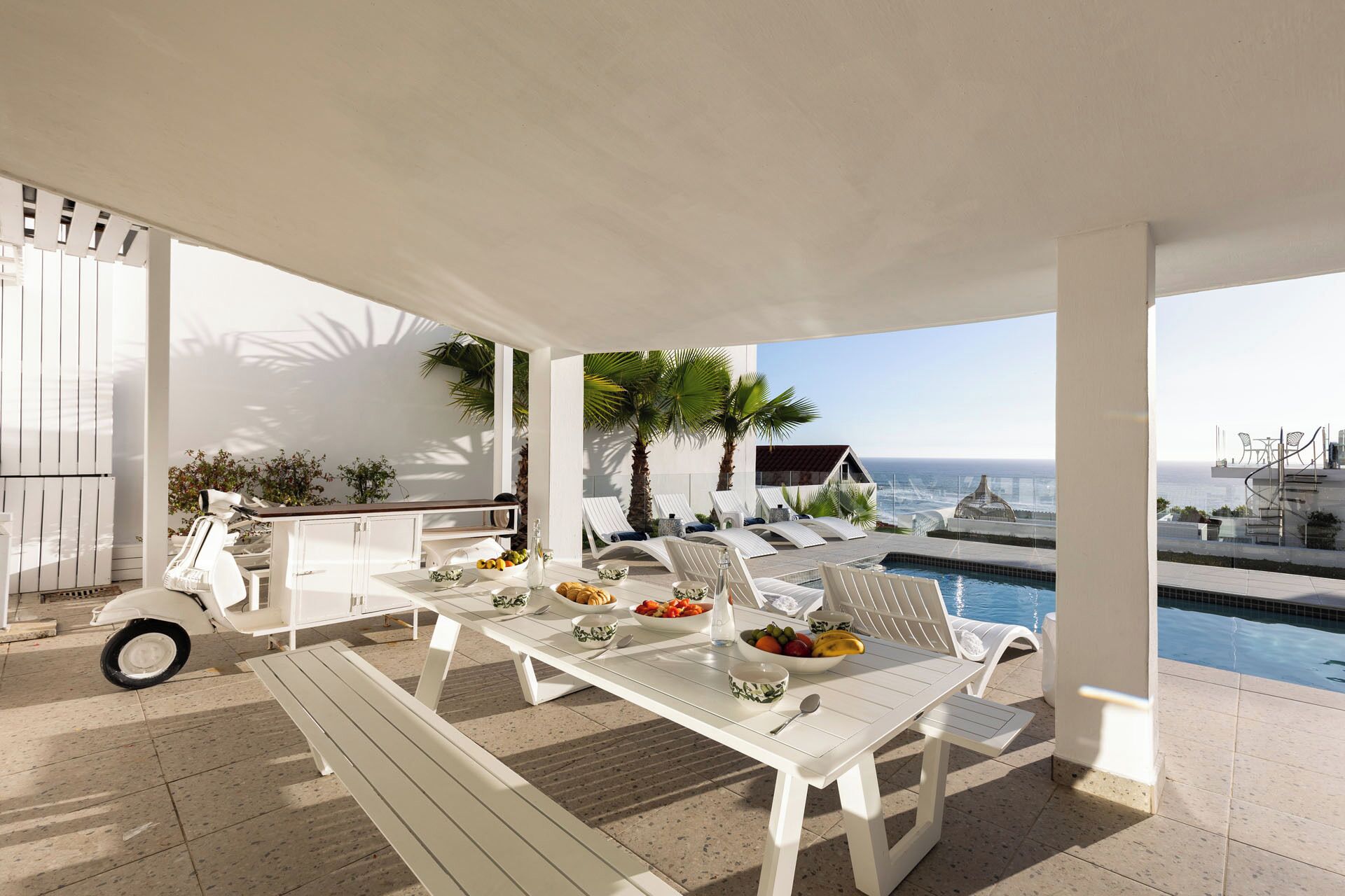 A covered outdoor dining area with a pool and lounge chairs, overlooking the sea.