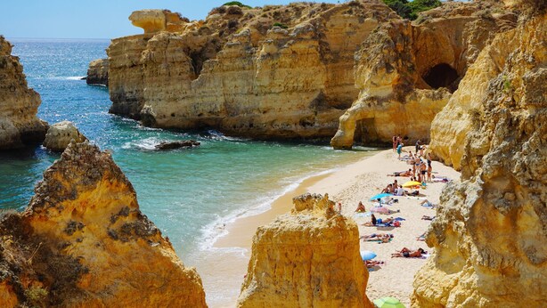 Une plage côtière bordée de falaises rocheuses, avec une eau d’un bleu limpide et des gens qui se prélassent au soleil.