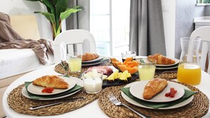 Une table de petit-déjeuner avec des croissants, des fruits et du jus, installée dans une pièce lumineuse agrémentée d’une plante et offrant une vue sur l’extérieur.