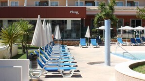 Un hôtel doté d’un espace piscine, de chaises longues, de parasols et d’un bâtiment avec de nombreux balcons.