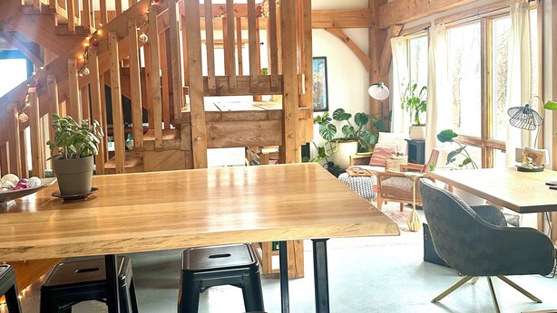 A spacious room with a wooden staircase, a dining table with chairs, and large windows with curtains.